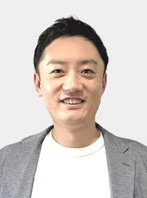 白いTシャツの上にグレーのジャケットを羽織った男性が、優しい笑顔でカメラを見ているポートレート写真です。清潔感のある明るい印象を与えます。