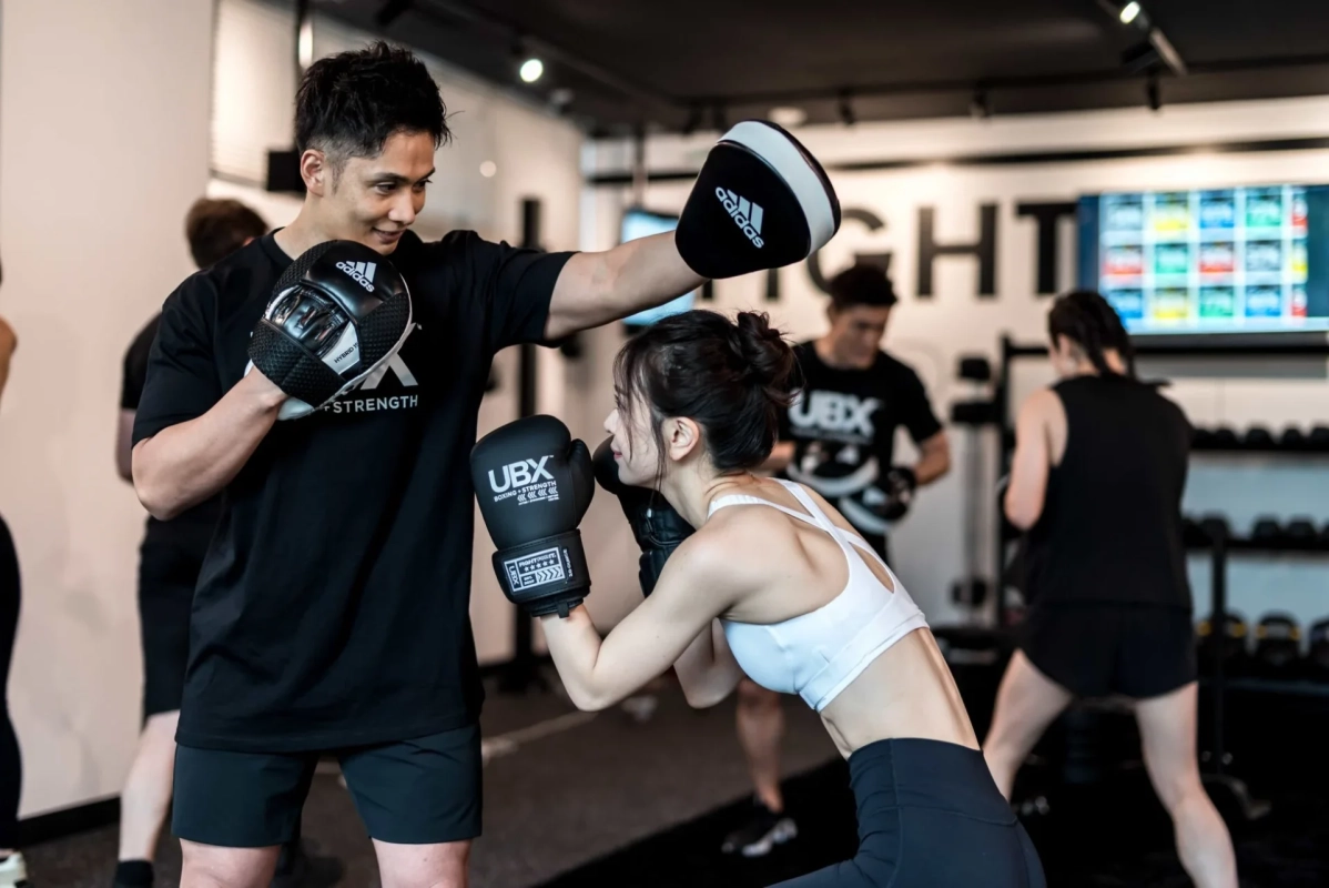 ジムで男性トレーナーがミットを持ち、女性がボクシンググローブをつけてパンチの練習をしている様子です。他の人々もトレーニングしており、活気あるフィットネス環境が伺えます。