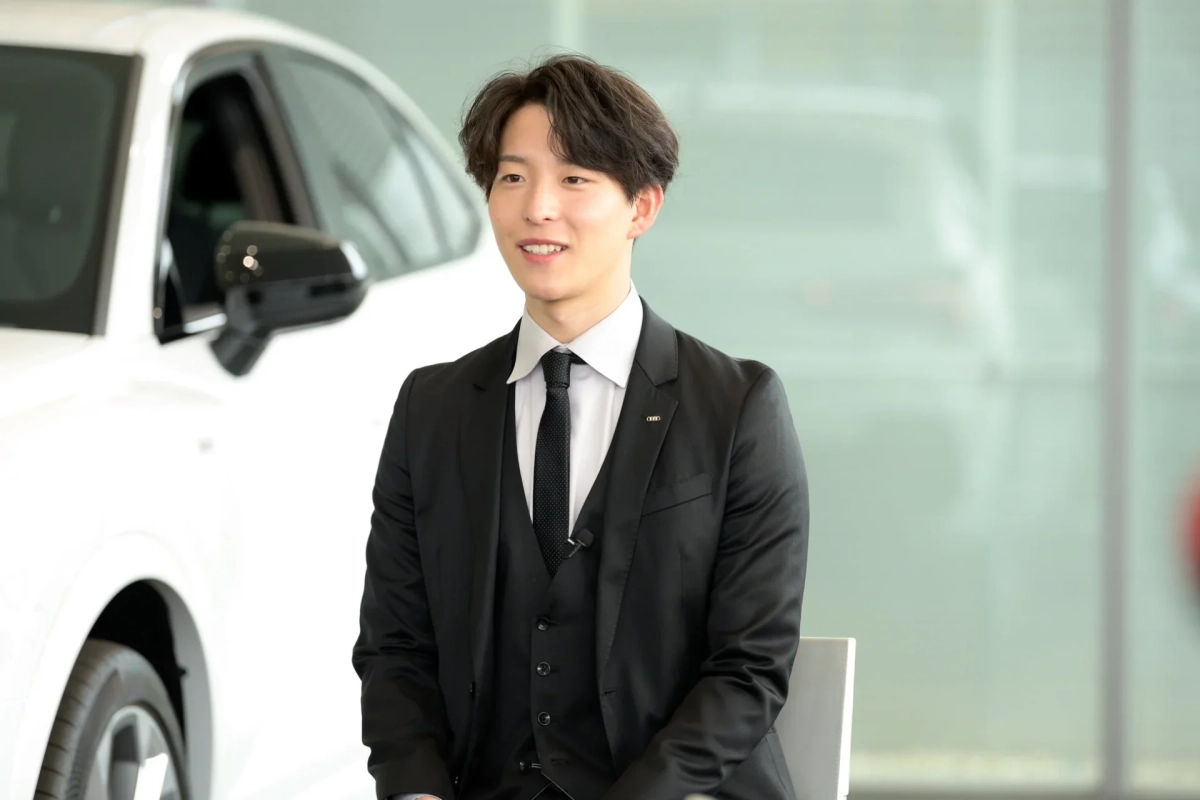 白い車を背景に、スーツを着た若い男性が笑顔で座っている様子です。