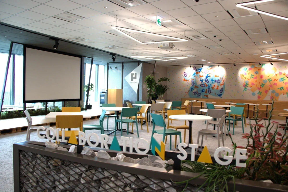 カラフルな家具が配置されたモダンで開放的なコラボレーションスペース。大きなスクリーンやアート性の高い壁画があり、「COLLABORATION STAGE」のサインが目を引く。会議やワークショップに適した多目的空間。