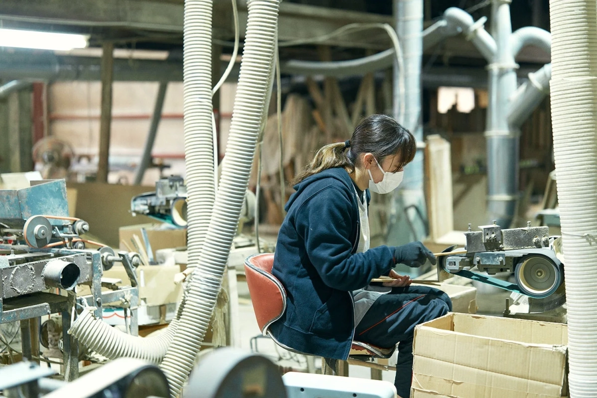 工場内でマスクを着用した女性が、ベルトサンダーのような機械を使って木材を加工する作業をしている様子