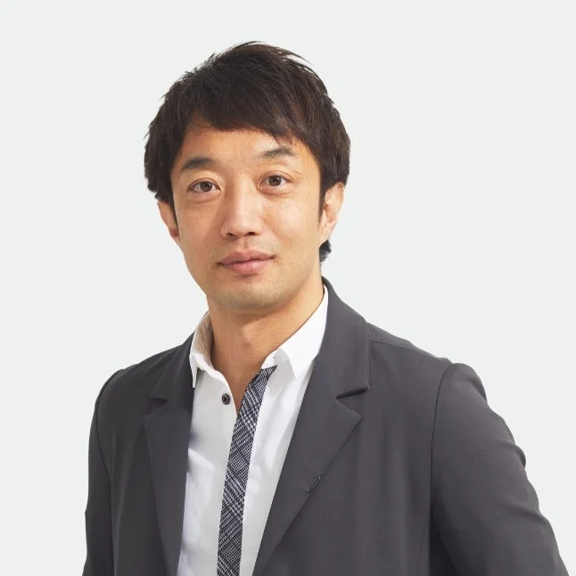 白い背景をバックに、ダークグレーのスーツジャケットと白いシャツを着用したアジア人男性のポートレートです。男性はカメラをまっすぐ見ており、真面目な表情をしています。