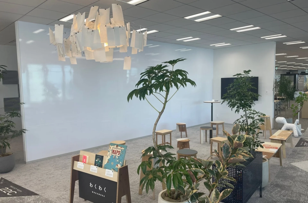 観葉植物が豊富に配置された、明るくモダンなオフィスラウンジです。白い壁とデザイン性の高い照明が特徴で、様々なスツールや本棚があり、リラックスできる創造的な空間が広がっています。