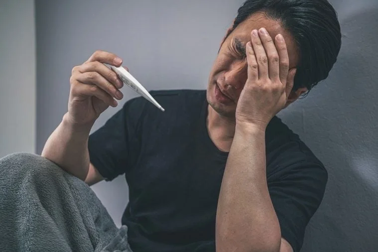 体調が悪そうな男性が体温計を持ち、顔を手で覆っています。発熱や病気で辛い状況を表しているようです。