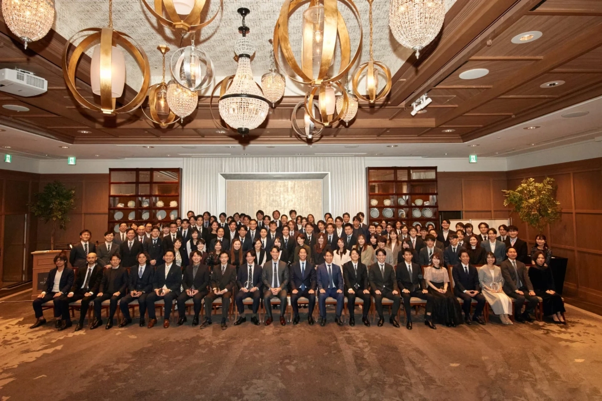 豪華なシャンデリアのある広々とした会場で、ビジネススーツやドレスを着用した大勢の人々が並んで撮影された集合写真