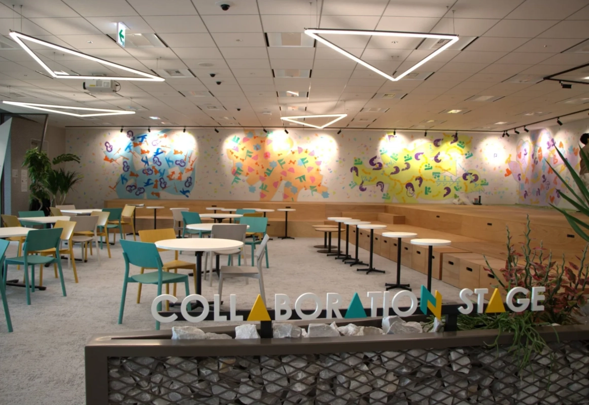モダンでカラフルな内装のコラボレーションスペースです。複数のテーブルと椅子、そして段差のあるステージ席が配置されており、「COLLABORATION STAGE」のサインが目立ちます。交流やイベントに適した多目的な空間です。