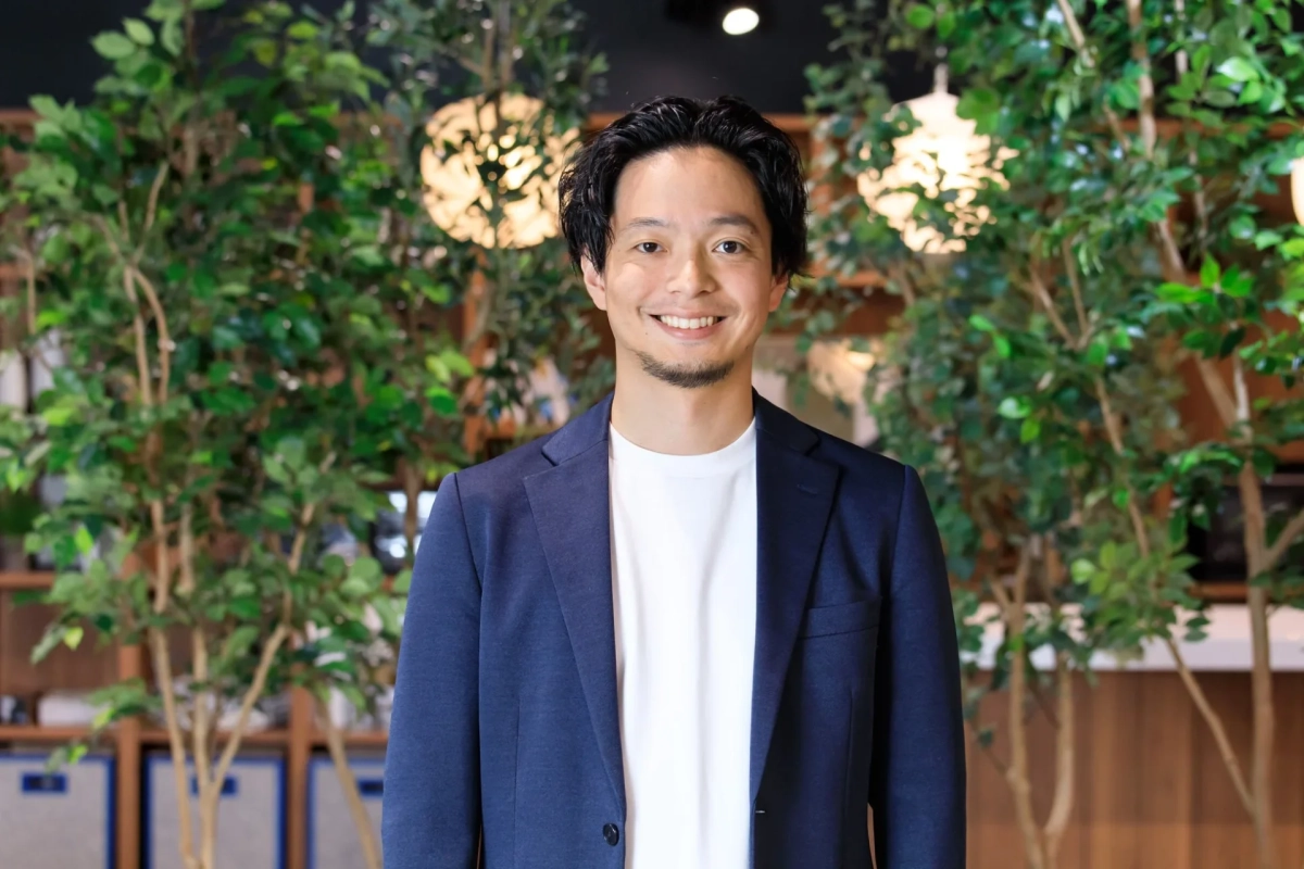 青いジャケットと白いTシャツを着た男性が、緑の植物と柔らかな照明を背景に笑顔でカメラを見ているポートレート写真