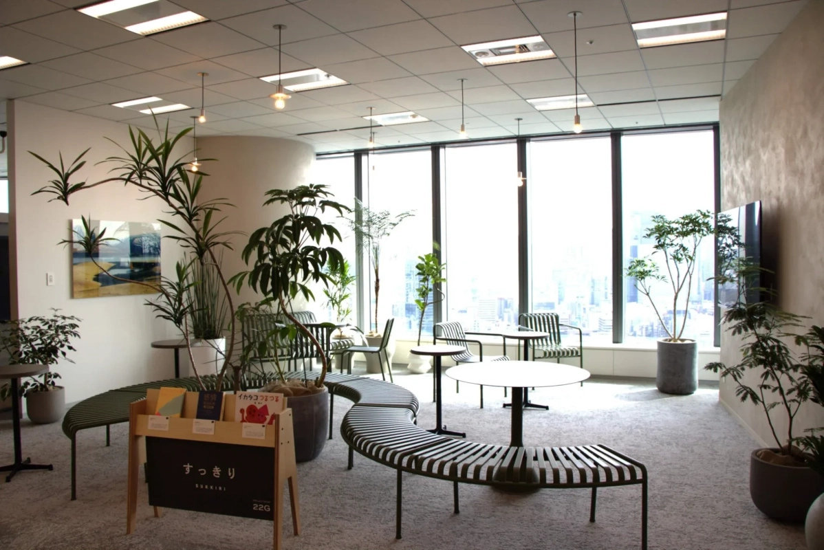 大きな窓から光が差し込む、広々としたモダンなオフィスラウンジです。多数の観葉植物とユニークなデザインのベンチやテーブルが配置され、リラックスできる快適な空間が広がっています。窓の外には都市の景色が望めます。