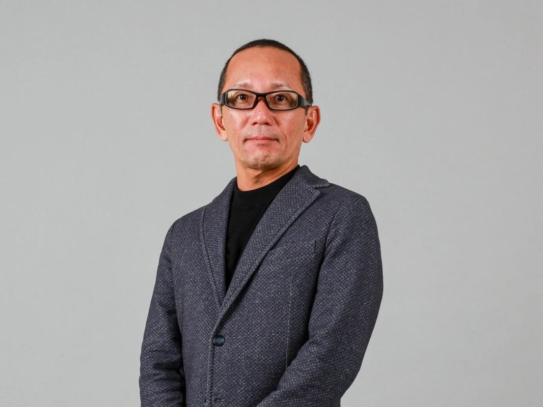 グレーの背景を背に、黒いタートルネックと紺色のジャケットを着用し、眼鏡をかけたアジア系の男性が正面を見ているポートレート写真です。