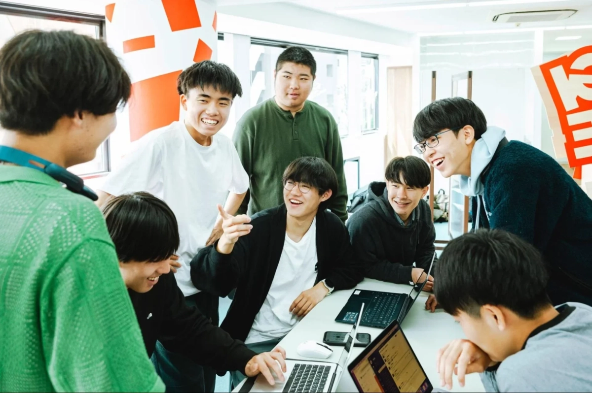 複数の若者がテーブルを囲み、ノートパソコンを使いながら笑顔で活発に交流している様子