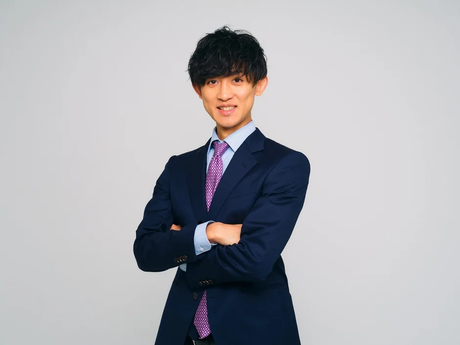 紺色のスーツと紫色のネクタイを着用した若い男性が、腕を組み笑顔で立っている