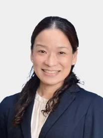 白い背景の前で、ダークスーツと白いブラウスを着用したアジア系の女性が笑顔で写っているプロフェッショナルな上半身のポートレートです。