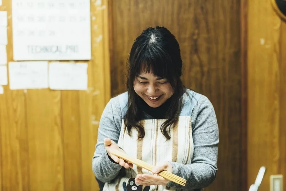 エプロンを着用した女性が、温かい笑顔で木製の細長い棒を両手で丁寧に扱っている様子