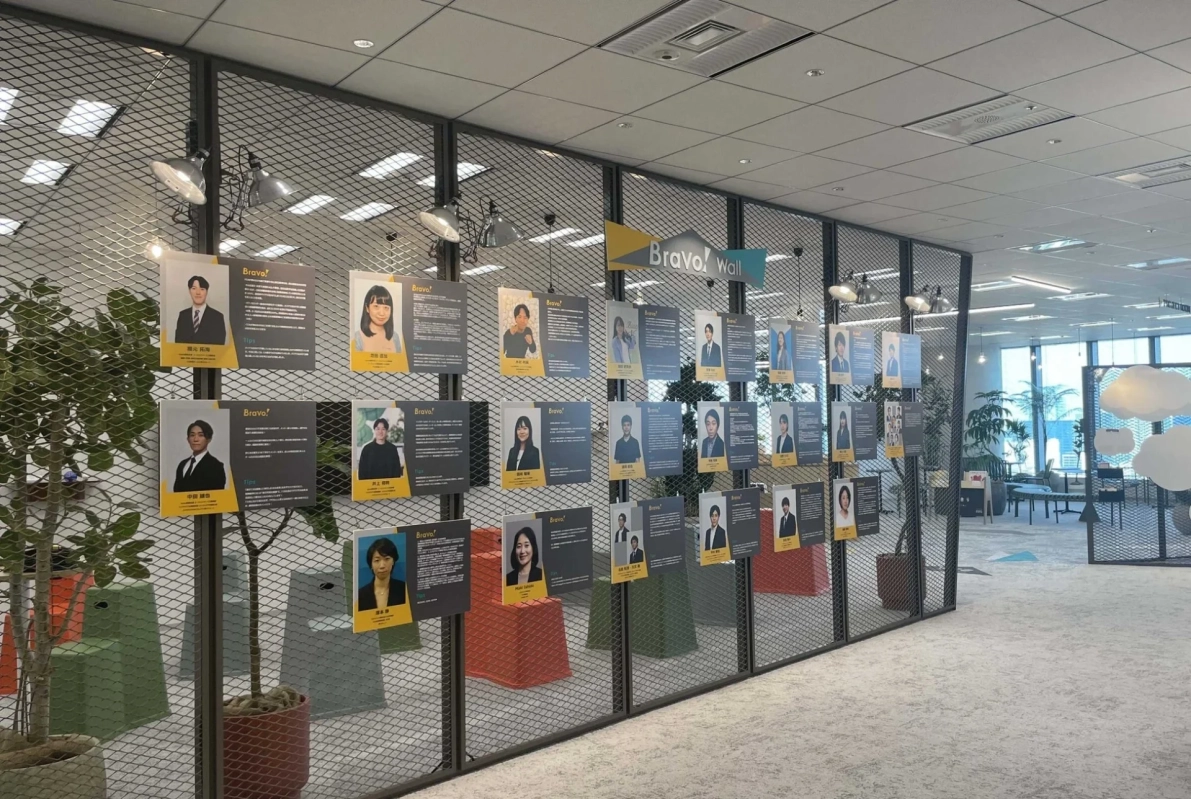 現代的なオフィス空間に設置された「Bravo! Wall」と呼ばれる社員紹介ボード。多数の社員の顔写真とプロフィールが展示されており、従業員への称賛や企業文化を示す壁面ディスプレイの様子を捉えている。