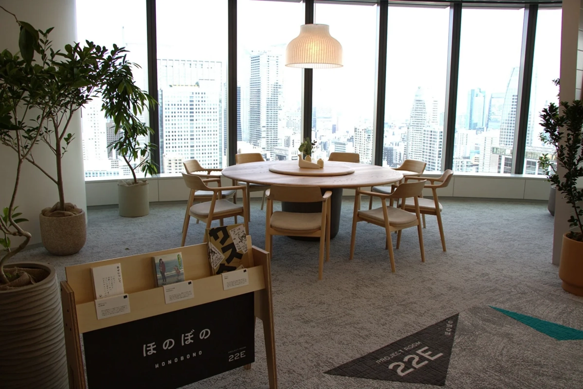高層ビルの窓から都市の景色が広がる、明るく広々としたモダンな会議室。木製の円卓と椅子、観葉植物が配置され、リラックスできる空間を演出。手前には「ほのぼの」と書かれた書籍棚が見えます。