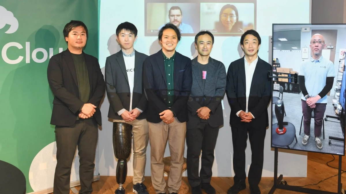 ビジネスイベントで撮影されたグループ写真。登壇者と思われる5人の男性が並び、背景のスクリーンにはオンライン参加者や義足と関連する製品が映し出されています。