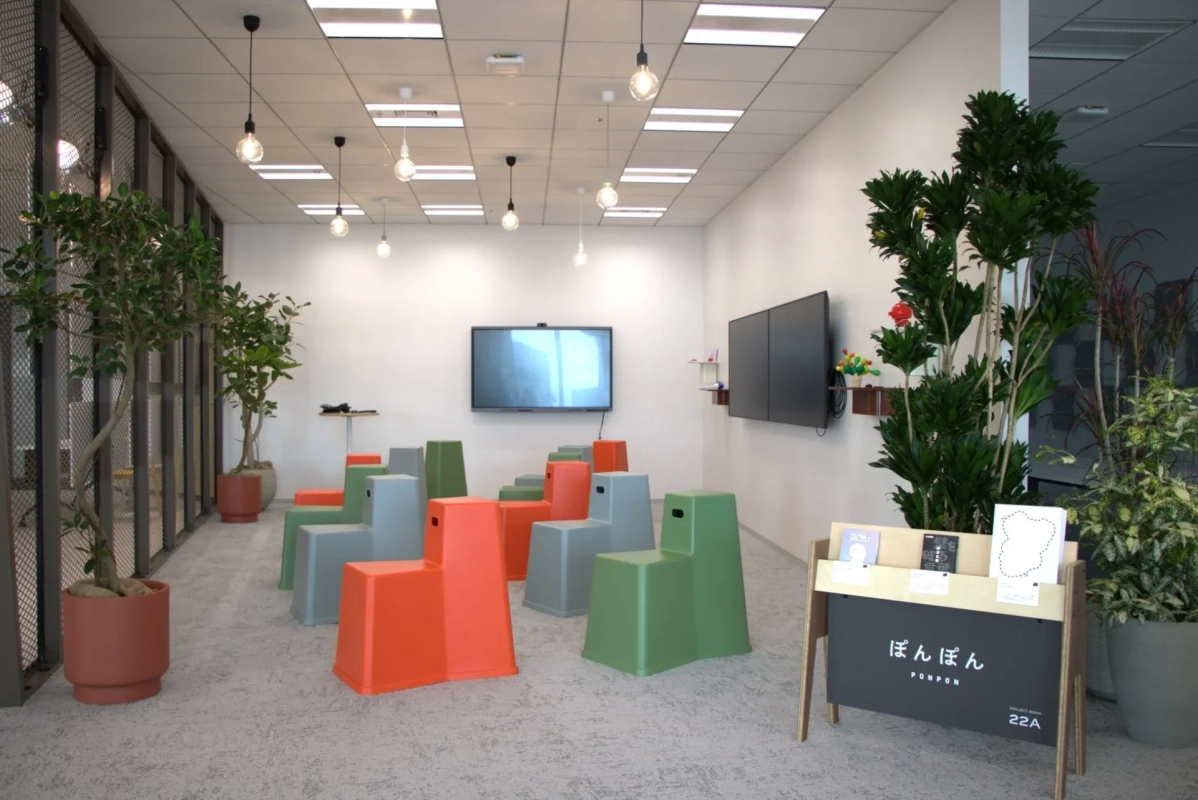モダンなデザインのオフィス共有スペースで、カラフルなステップ状の椅子が複数並べられています。壁には大型ディスプレイが設置され、観葉植物やデザイン性の高い照明が配されており、会議や休憩に利用できる多目的空間です。