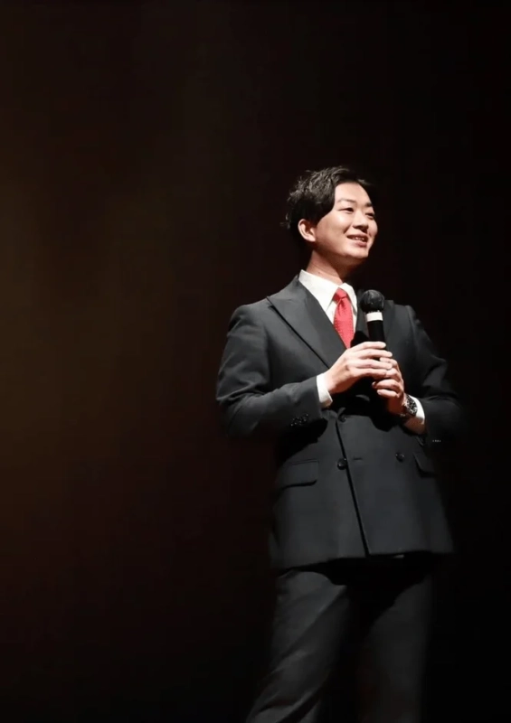 黒いダブルスーツに赤いネクタイを着用した男性がマイクを持ち、笑顔で何かを話している