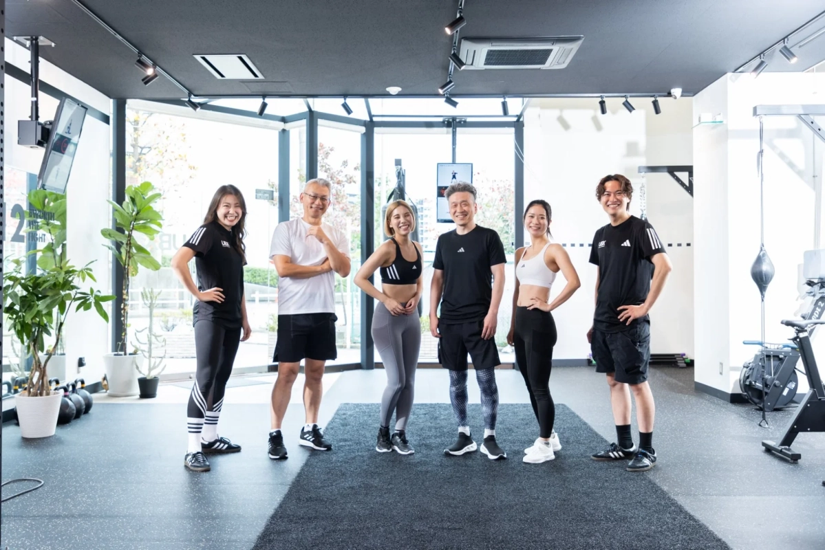モダンなジムで、様々な年齢層の男女6人がスポーツウェアを着用し、笑顔でカメラにポーズをとっています。健康的なライフスタイルと活気ある雰囲気が伝わる一枚です。