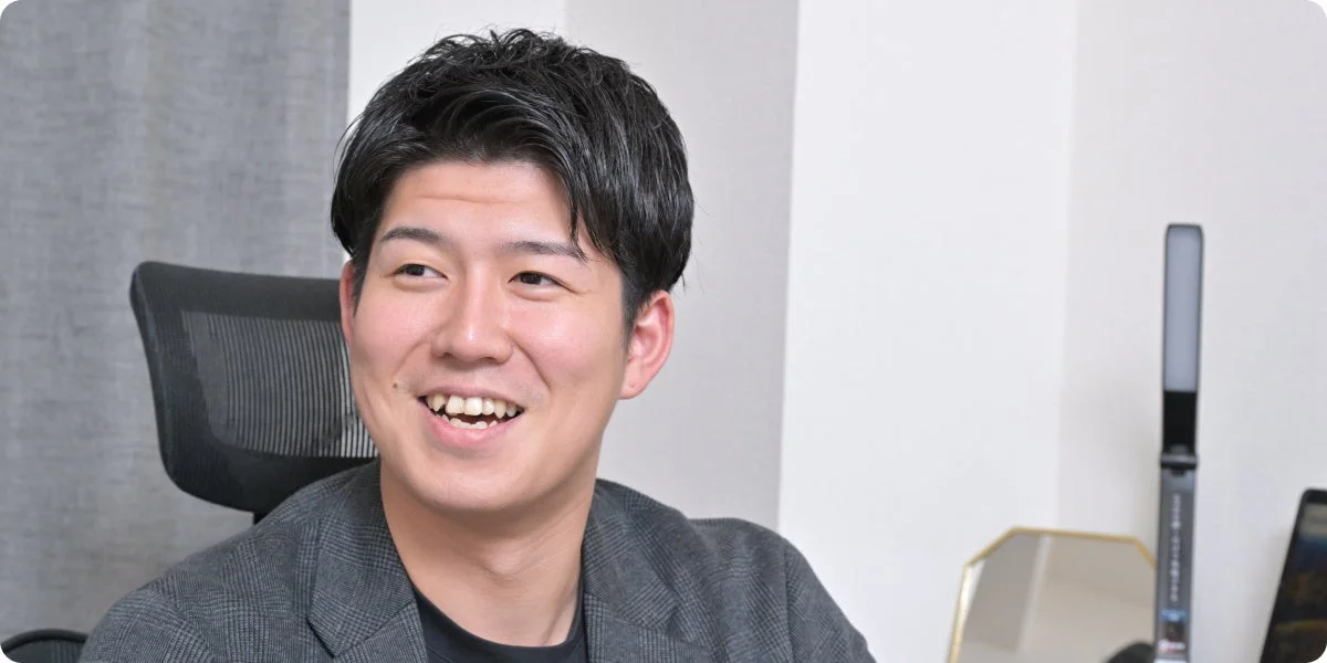 笑顔で左方向を見ている若い男性のポートレート。オフィスのような空間で、カジュアルなジャケットを着用し、リラックスした表情を見せている。背景にはデスクライトや鏡が見える。