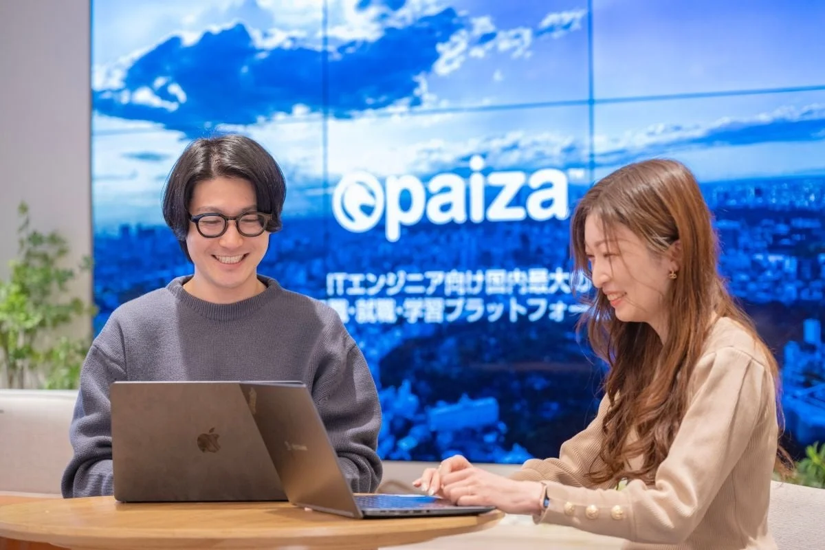 男女2人が笑顔でノートパソコンに向かっている様子。背景には「paiza ITエンジニア向け国内最大級の転職・就職・学習プラットフォーム」というロゴと文字が表示されており、IT関連の仕事や学習に取り組むポジティブな雰囲気が伝わります。