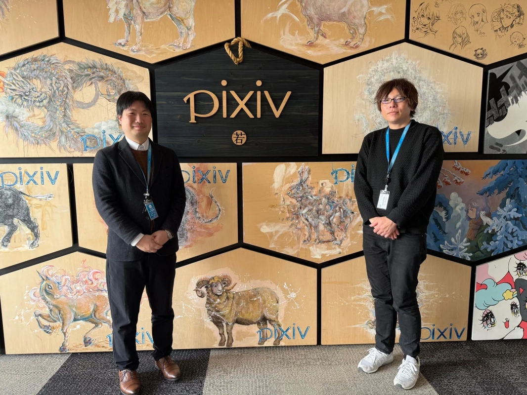 Pixivのロゴと動物のイラストが描かれた壁の前で、2人の男性が立っています。