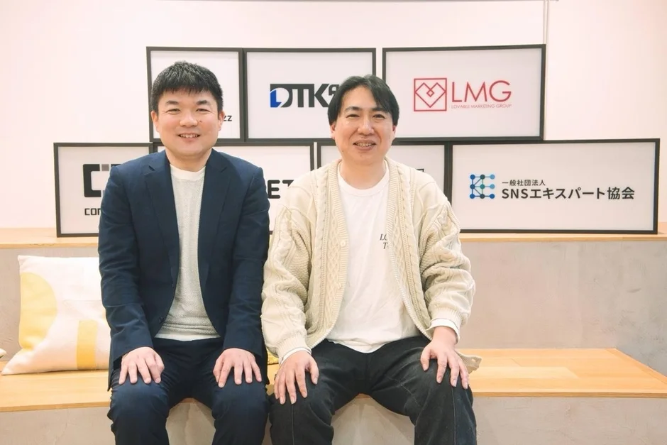 笑顔の男性2人が、複数の企業や団体のロゴが飾られた壁を背景に座っているビジネスシーンの写真。