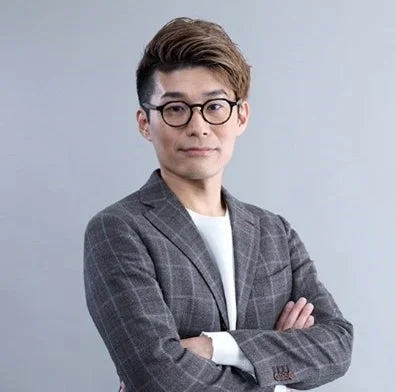 眼鏡をかけたアジア人男性が、グレーのチェック柄スーツを着用し、腕を組んでカメラを見つめているポートレートです。シンプルな背景で、ビジネスシーンやプロフィール写真に適した落ち着いた印象を与えます。