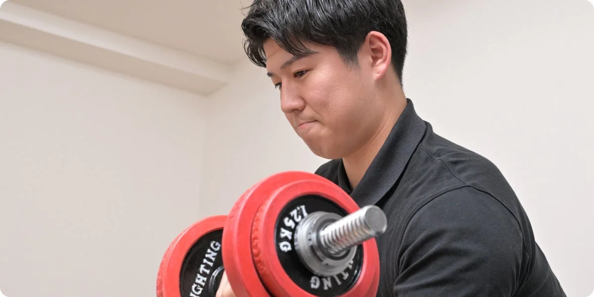 黒いポロシャツを着た若い男性が、赤いダンベルを使って筋力トレーニングに集中している様子を捉えた画像です。真剣な表情で重りを持ち上げています。