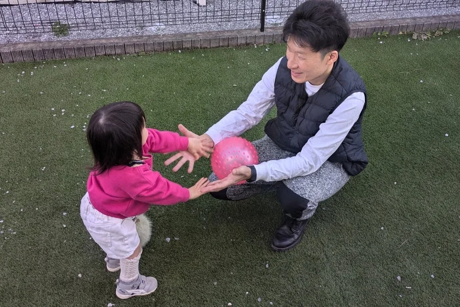 人工芝の上で、大人が子供にピンク色のボールを渡そうとしている瞬間の写真です。二人は笑顔で、楽しそうに交流しています。