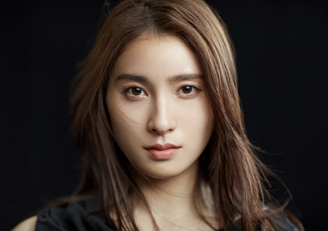 暗い背景をバックに、長い茶色の髪の若い女性がカメラをまっすぐに見つめているポートレート。