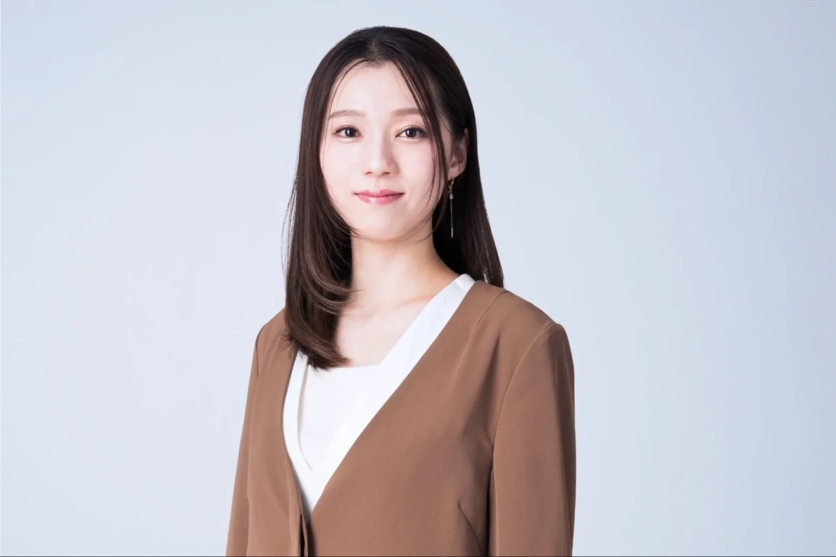 明るい背景をバックに、長い黒髪の若い女性が優しく微笑んでいます。茶色のジャケットと白いインナーを着用し、カメラに穏やかな表情を向けている上半身のポートレートです。