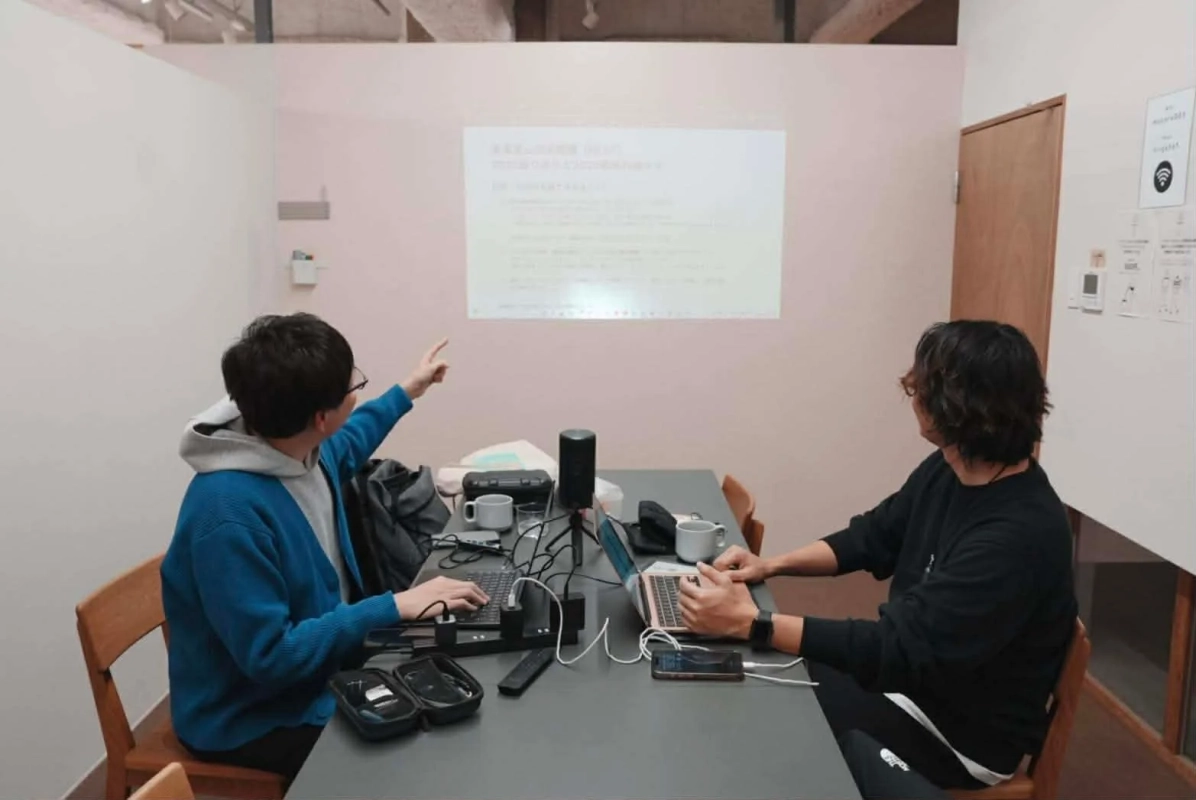 オフィスのような空間で、2人の男性がプロジェクターに映された資料を見ながらラップトップで作業している様子