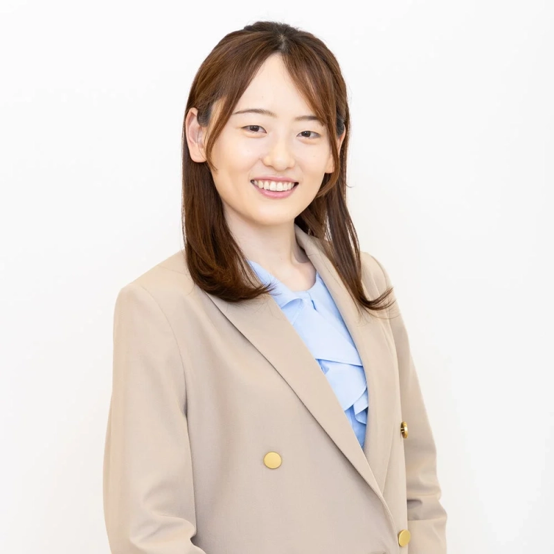 白い背景の前で、ベージュのジャケットと水色のブラウスを着用した若い女性が笑顔で立っているポートレート写真です。