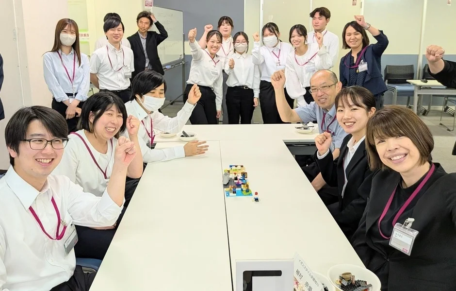 オフィスまたは会議室で、多くの人々が笑顔でガッツポーズをしている集合写真。テーブルにはレゴブロックがあり、チームビルディングやワークショップの活気ある様子が伝わります。