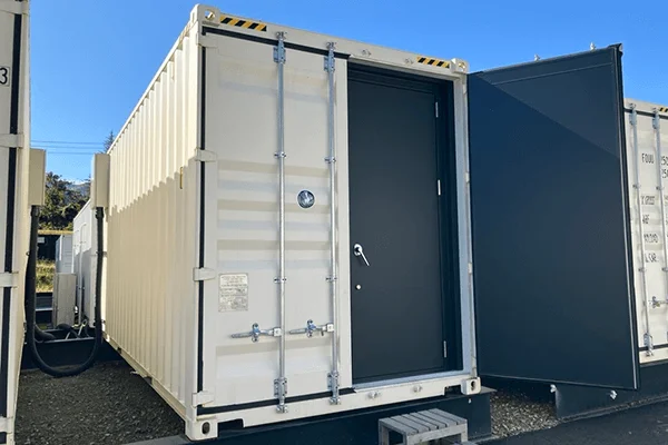 白い輸送コンテナが複数並んでいます。手前のコンテナには黒い扉が取り付けられており、何らかの設備が収納されているか、あるいは作業スペースとして利用されているようです。晴れた日の風景です。