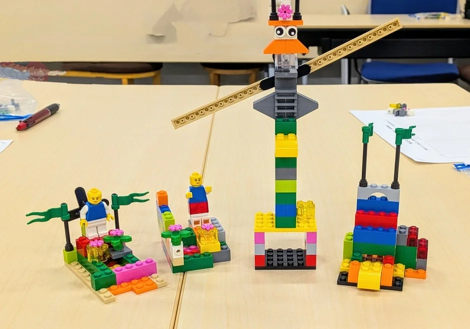 木製のテーブルの上に、様々な色と形のレゴブロックで作られた4つの作品が並んでいます。クレーンのような高い構造物や、ミニフィギュアが乗った小さな台座など、子供たちの創造性が光る組み立ておもちゃの様子を捉えた画像です。