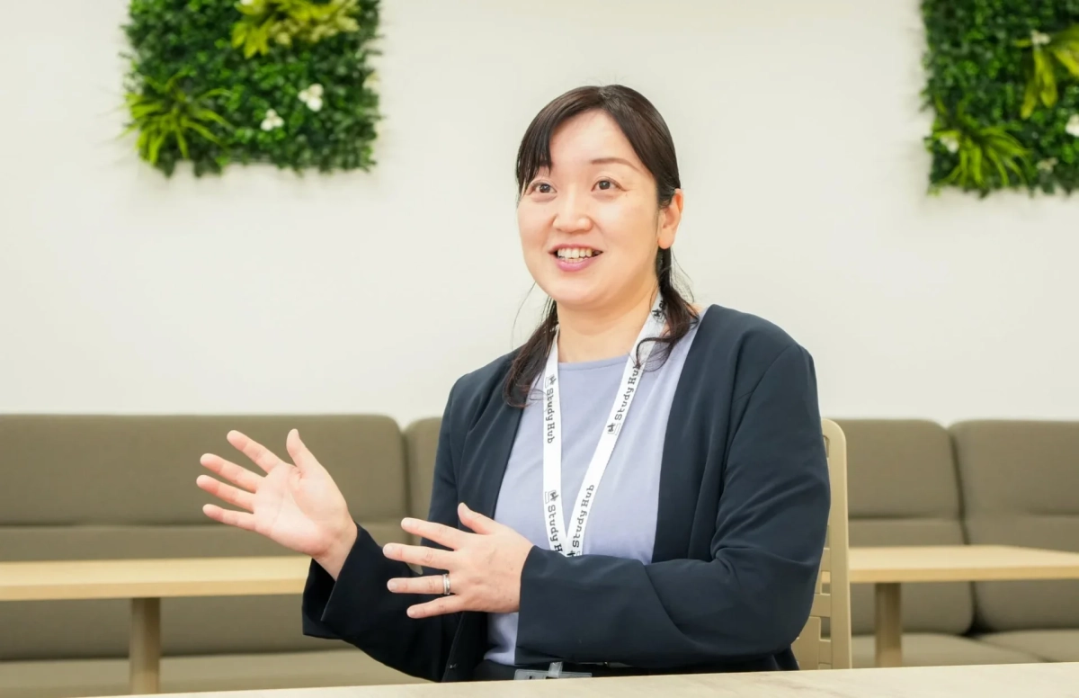 オフィスのような空間で、Study Hubのランヤードを着用したアジア人女性が笑顔で身振り手振りを交えながら話している様子です。背景には緑の装飾が施されています。