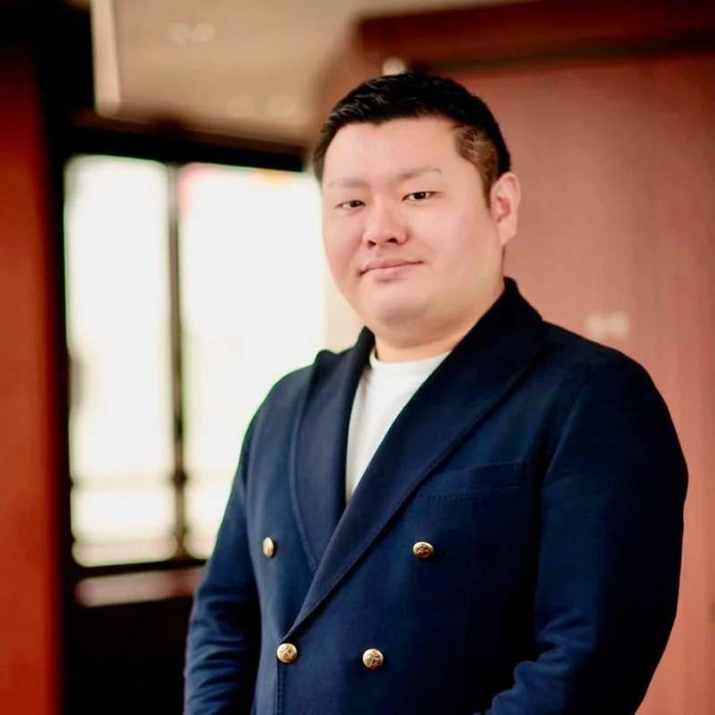 紺色のダブルブレストジャケットを着たアジア人男性のポートレートです。金色のボタンが特徴的で、白いTシャツと合わせています。室内でカメラをまっすぐ見つめる彼の表情からは、落ち着きと自信が感じられます。