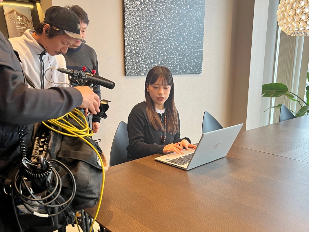 会議室のような空間で女性がノートパソコンで作業している様子を2人の男性が映像機材を使って撮影しているシーン