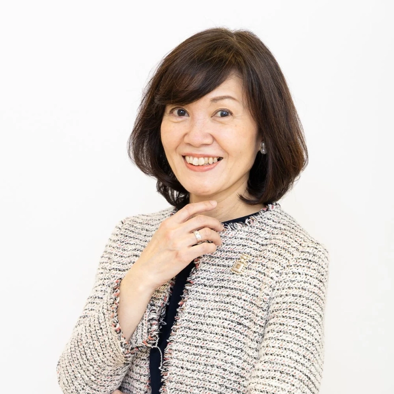 白い背景の前で、カメラに笑顔を向ける日本人女性のポートレート。ツイードジャケットを着用し、指輪とブローチを身につけており、明るくプロフェッショナルな印象を与えます。