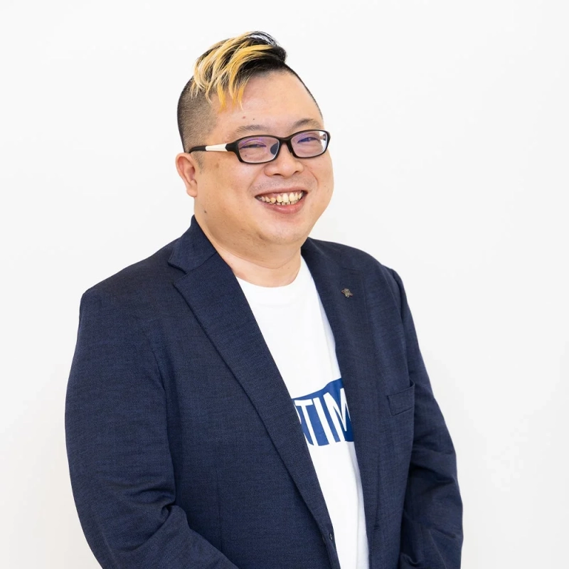 白い背景の前で笑顔を見せるアジア人男性のポートレート。彼は眼鏡をかけ、紺色のブレザーの下に白いTシャツを着ており、特徴的な金髪のモヒカンスタイルの髪型が目を引きます。