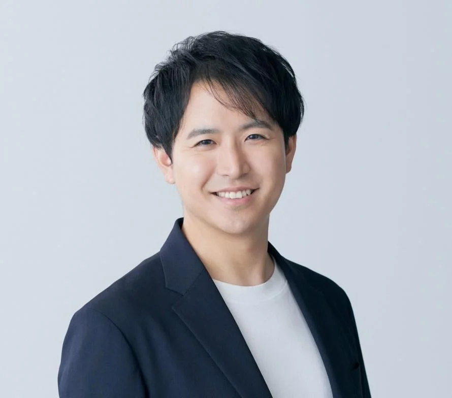 明るい背景の前で、ダークなジャケットと白いTシャツを着用したアジア人男性が、カメラに向かって優しく微笑んでいる上半身のポートレート写真です。