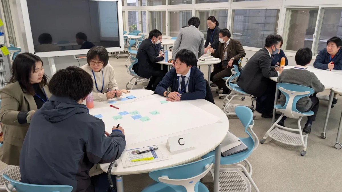 明るい部屋で、複数のグループに分かれた大人たちが円卓を囲んで議論やワークショップを行っている様子