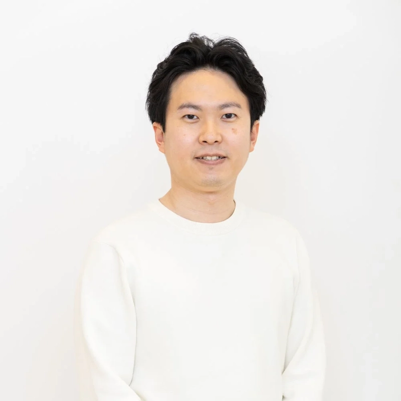 白い無地の背景を背に、白いセーターを着た日本人男性がカメラに向かってわずかに微笑んでいるポートレート写真です。