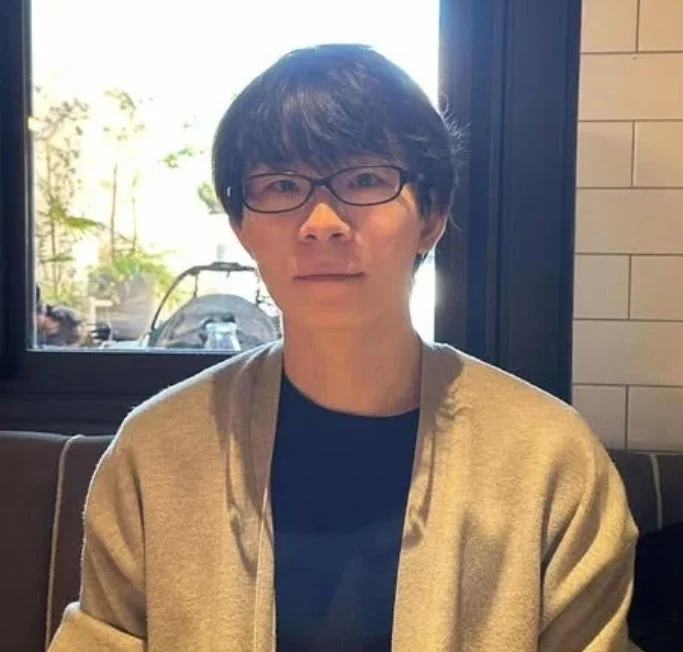 眼鏡をかけたアジア人男性がカメラを見つめているポートレートです。ベージュのカーディガンを着用し、背景には窓から見える緑と白いタイルがあります。