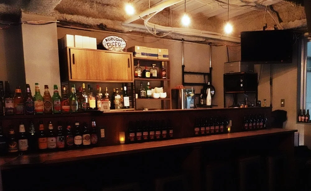 様々な種類のビールや洋酒が並べられたバーカウンターの様子です。温かみのある照明で照らされ、落ち着いた雰囲気が漂う店内の様子がうかがえます。