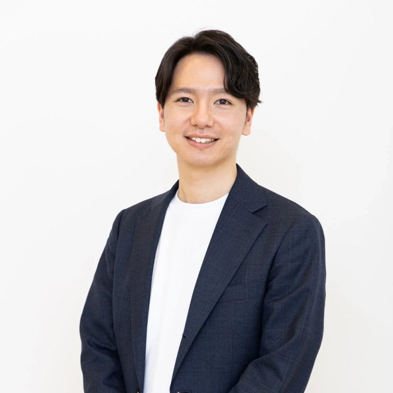 白いTシャツの上にダークスーツを着用した若いアジア人男性が、カメラに向かって優しく微笑んでいる上半身のポートレートです。