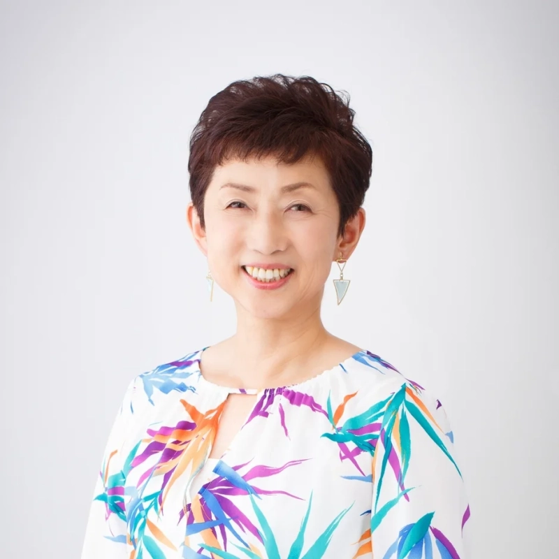 白い背景の前で、カラフルな葉柄のトップスを着用したショートヘアの日本人女性が、明るく親しみやすい笑顔を見せているポートレート写真です。