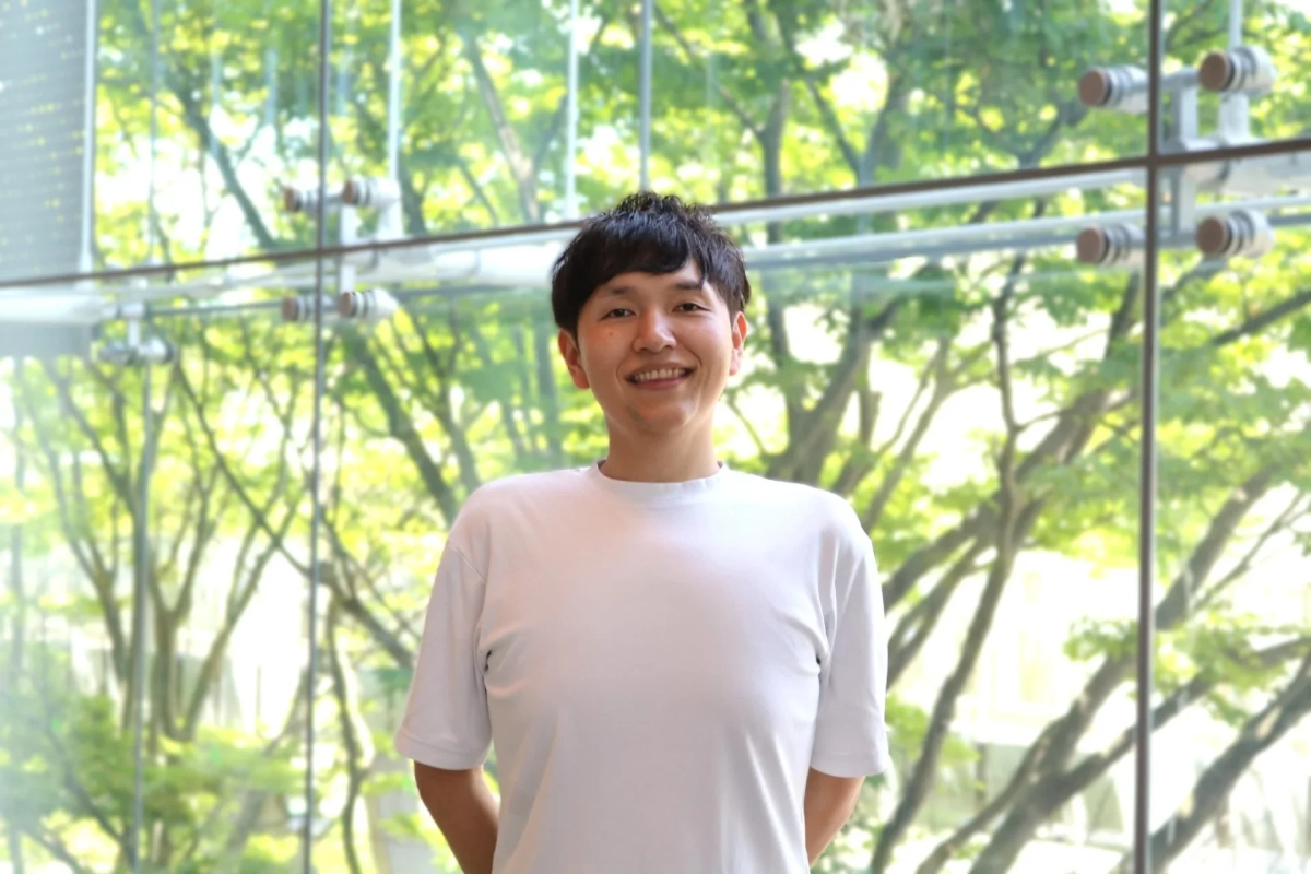 白いTシャツを着た男性が、窓越しの緑豊かな木々を背景に笑顔で立っているポートレート